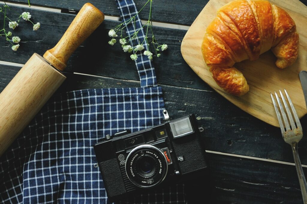 A wooden table displays croissants, a camera, and a knife, highlighting the art of creative food photography.