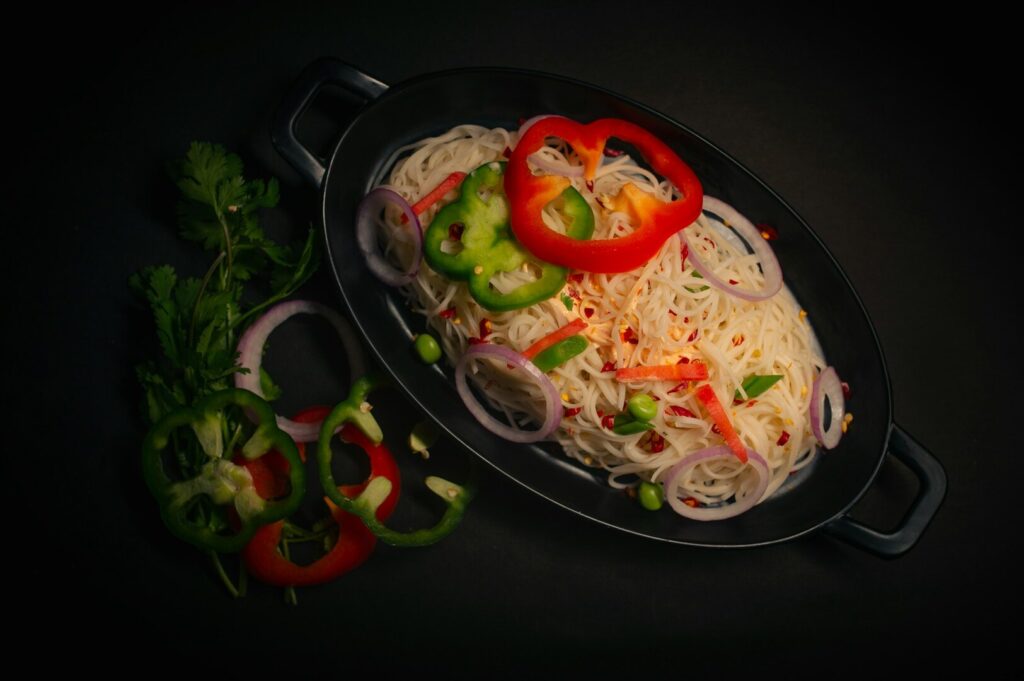 A close-up of a plate filled with noodles and peppers, artistically presented on a black background, highlighting Singaporean cuisine.