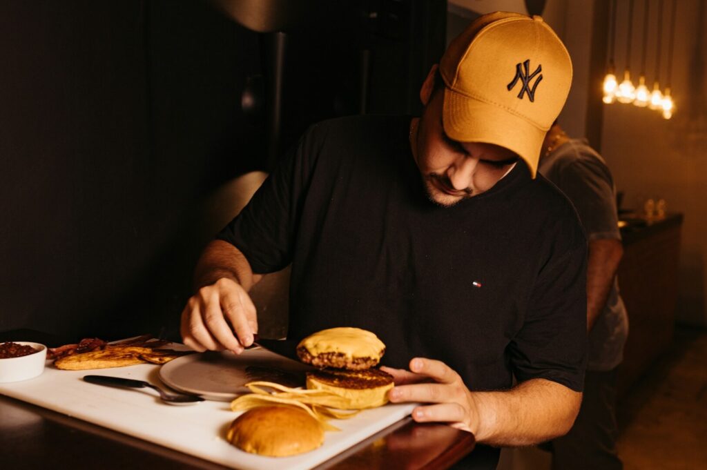 A man wearing a hat slices a burger, highlighting the art of professional food photography.