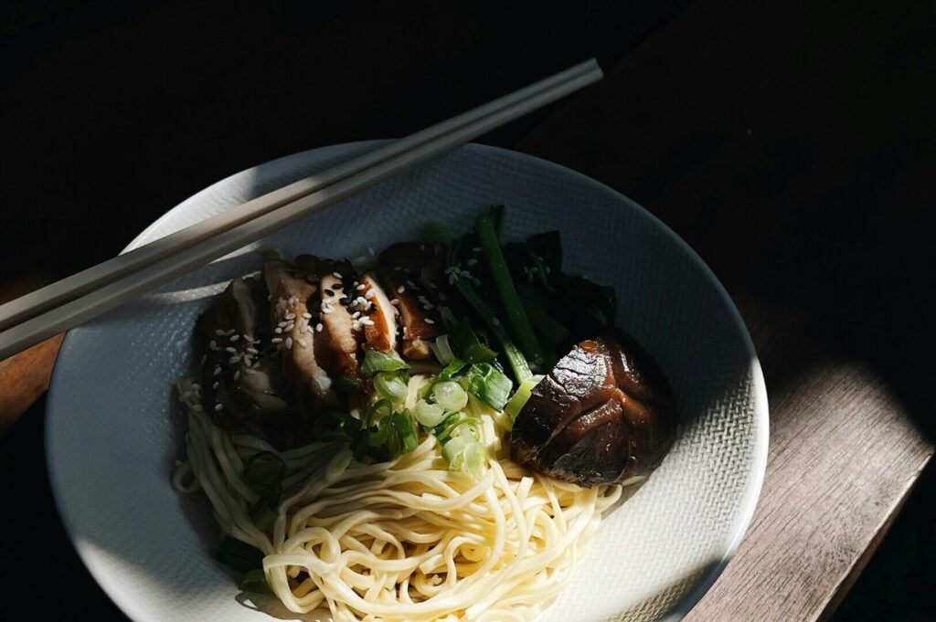 A white bowl of noodles and mixed vegetables, artfully arranged on a rustic table, showcasing vibrant food photography.