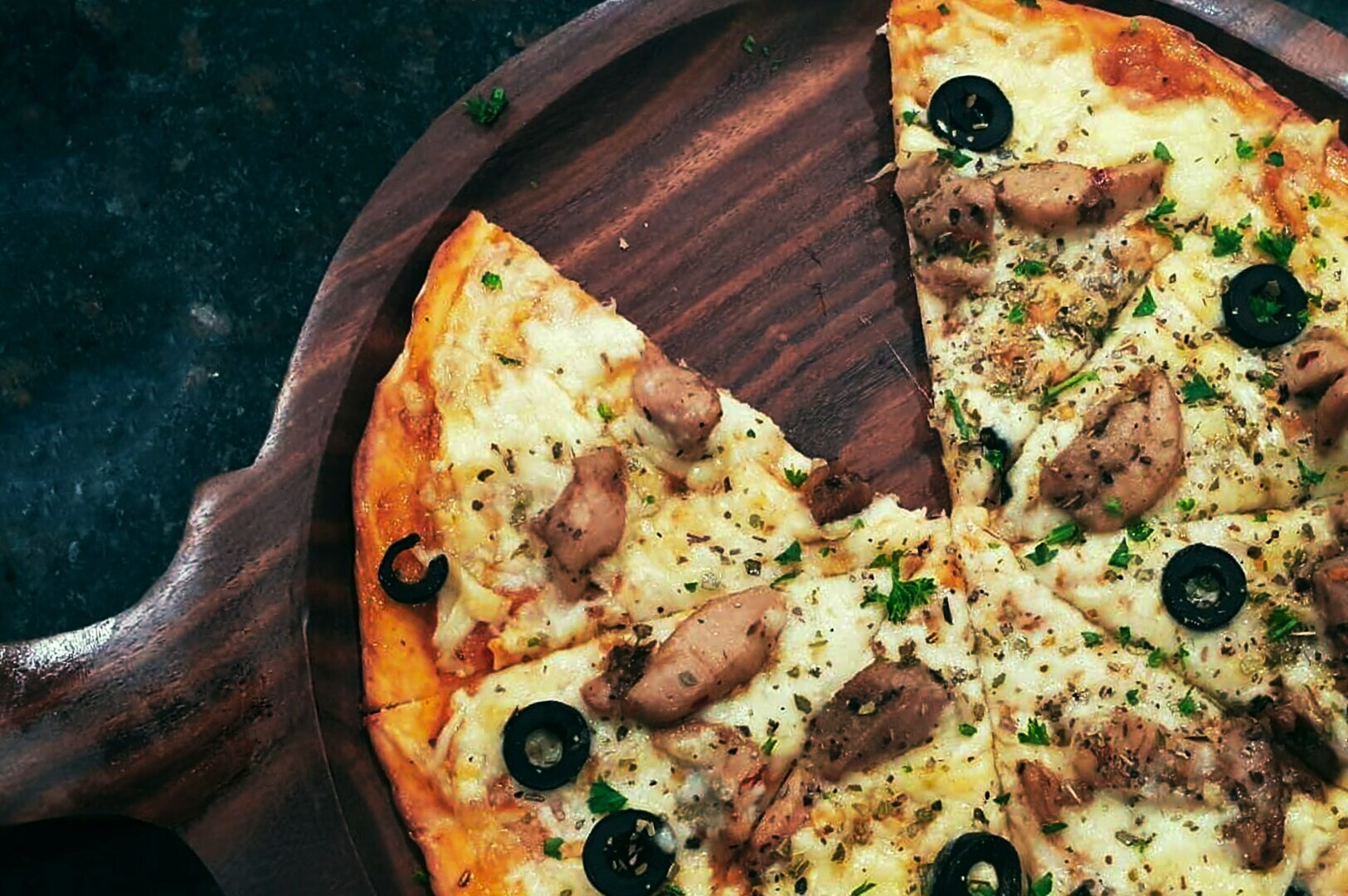 A wooden board displaying a sliced pizza topped with chicken, black olives, and herbs. The pizza's golden cheese and crust appear savory and appealing.