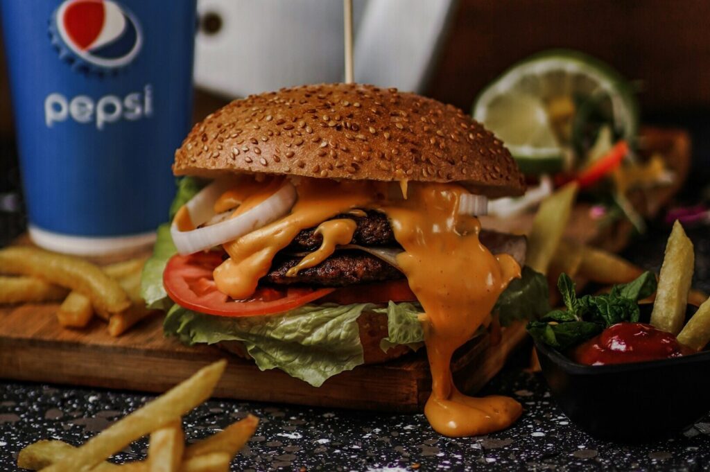 Close-up of a cheeseburger with lettuce, tomato, onion, and melted cheese on a sesame bun. Fries and a Pepsi cup in the background, creating a tasty setting.