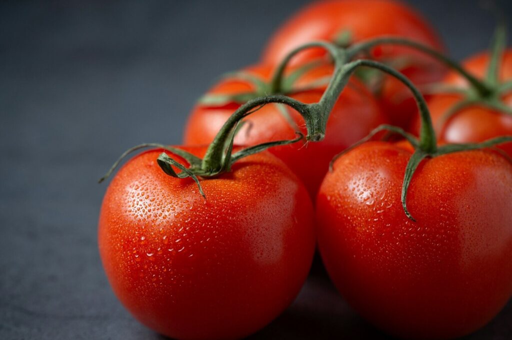 Fresh red tomatoes with dewdrops rest on a dark surface. They have green stems and are vibrant against the muted backdrop, suggesting freshness.