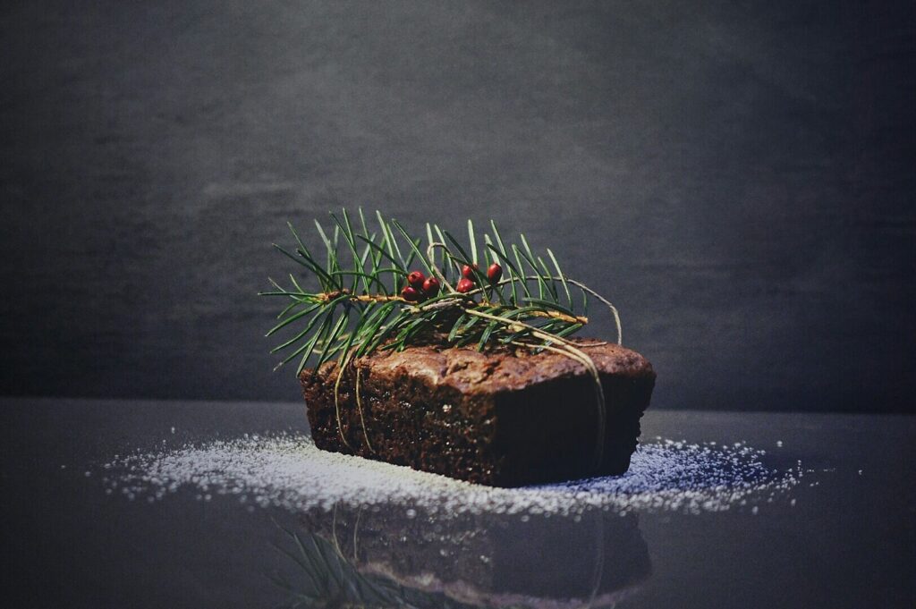 A festive brown loaf garnished with pine sprigs and red berries rests on a dark surface sprinkled with powdered sugar, conveying a rustic holiday feel.