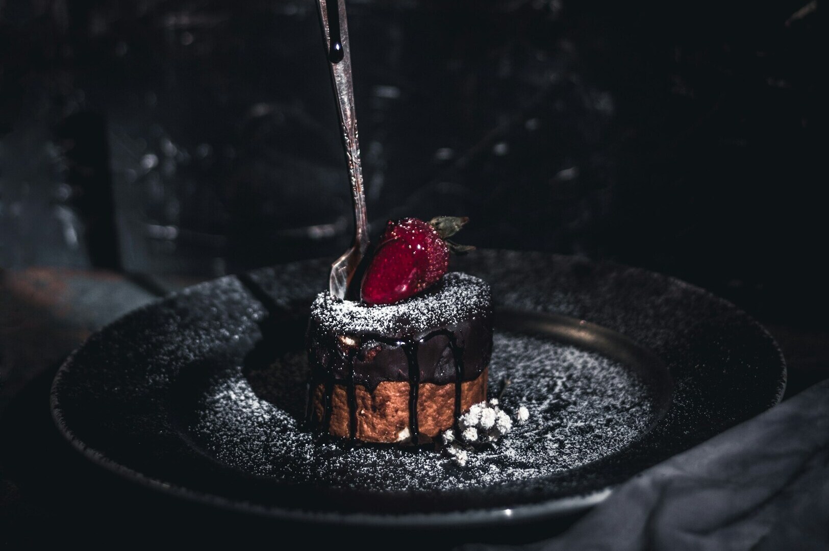 Decadent chocolate cake drizzled with syrup, topped with a bright red strawberry and sugar dusting, sits on a dark plate, set against a moody backdrop.