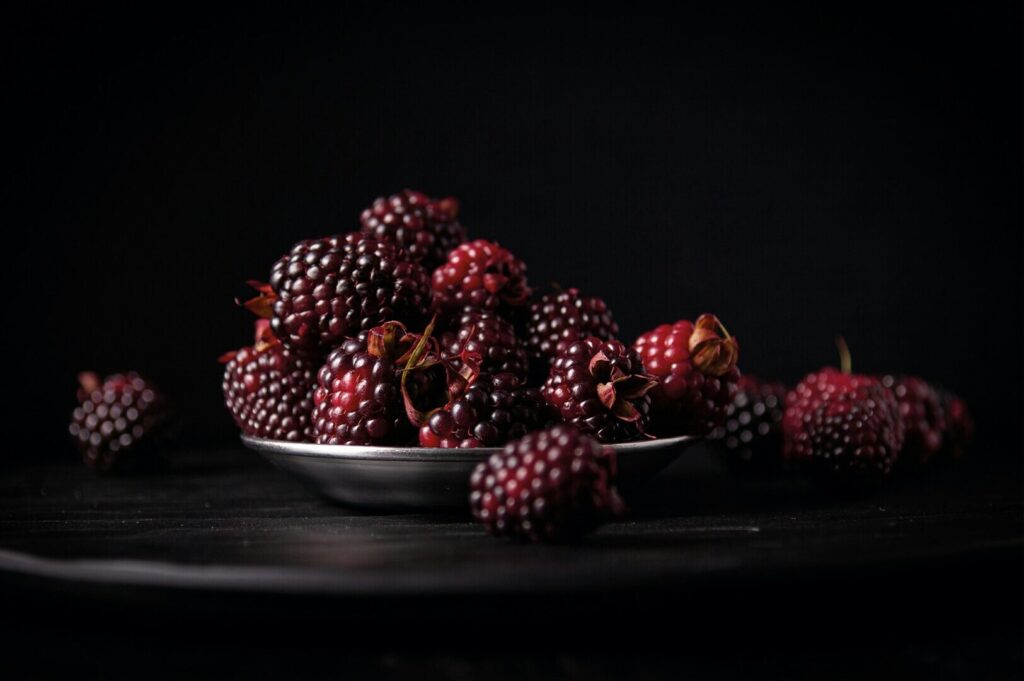 A pile of ripe blackberries sits in a bowl against a dark background. The berries are richly textured, with deep red hues, creating a moody, elegant tone.