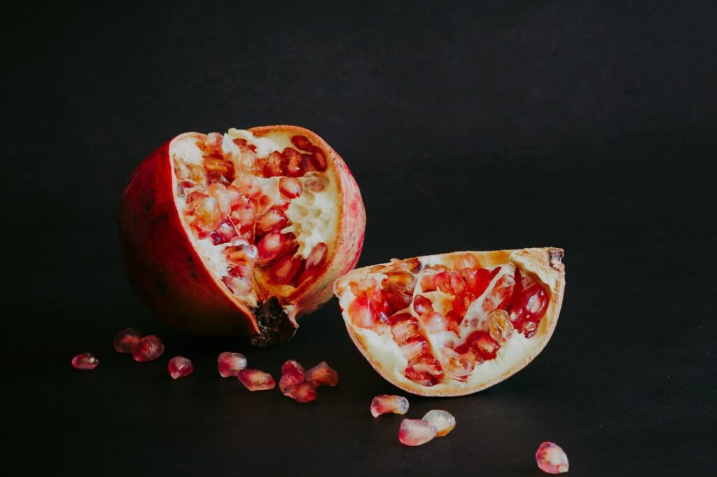 Half a pomegranate and scattered seeds on a dark background. The red seeds glisten, contrasting with the deep tones, conveying freshness and vibrancy.