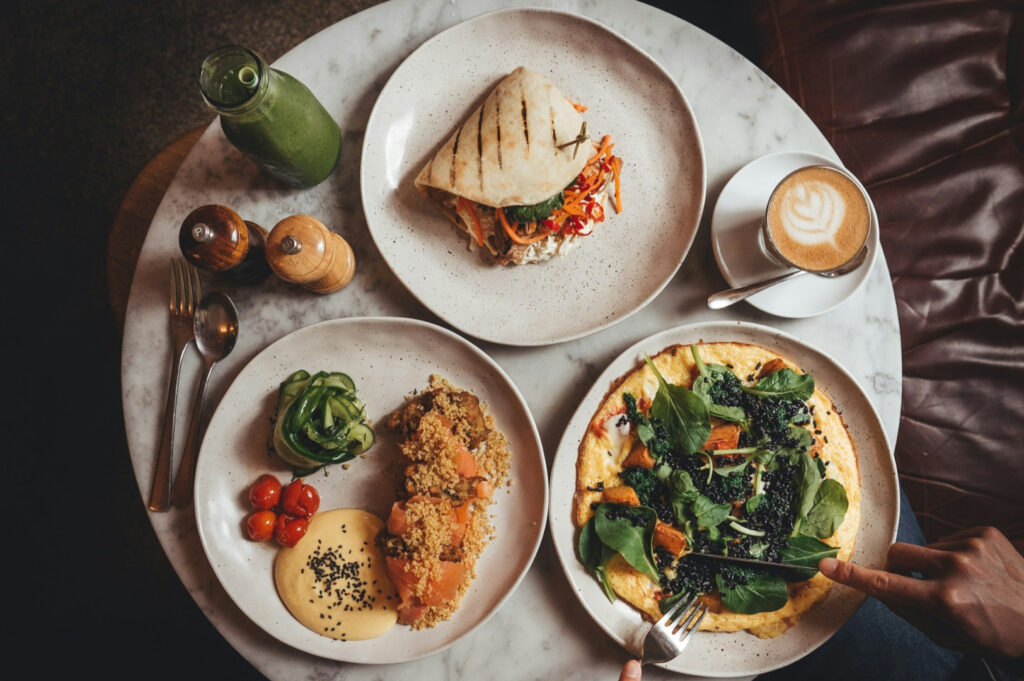 A round table with plates of diverse dishes: a grilled pita sandwich, a vibrant kale omelette, and crusted salmon. A smoothie and latte add a cozy feel.