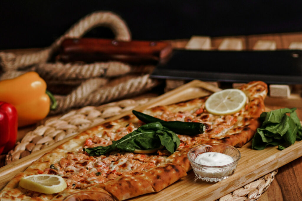 Flatbread topped with spinach, lemon slices, and green chilies on a wooden board. Nearby, a small bowl of sauce. Peppers and rope in the background.