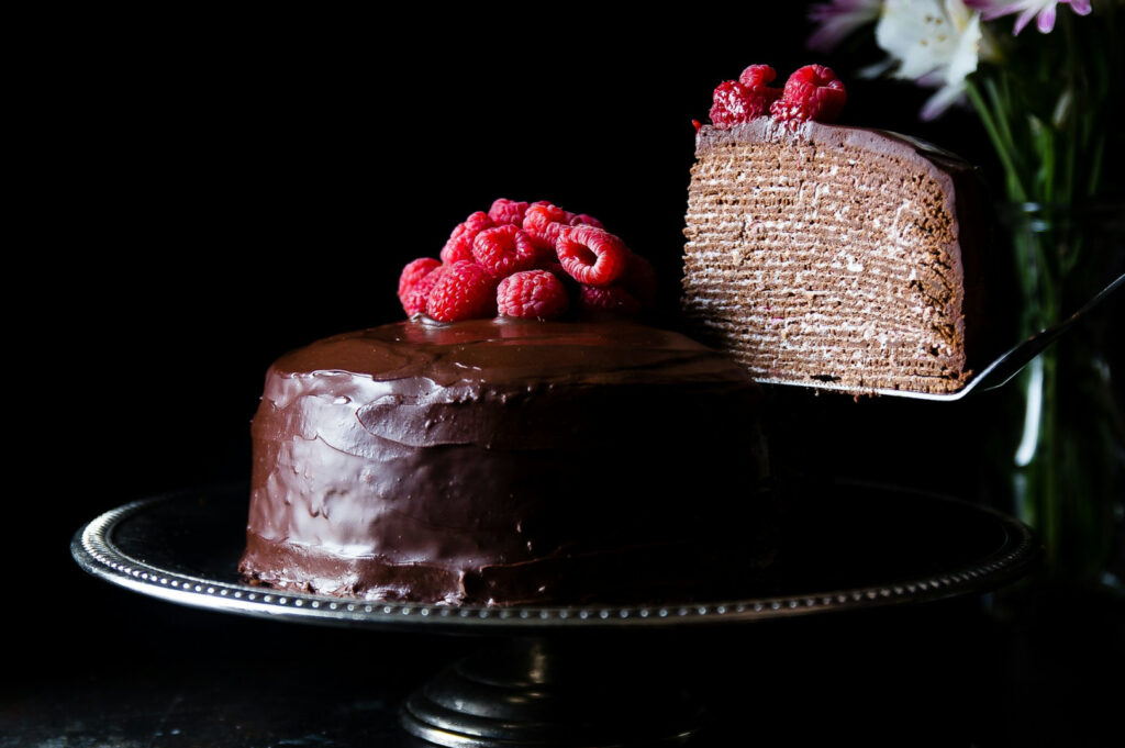 Chocolate cake with smooth frosting topped with fresh raspberries on an elegant stand. A slice is being lifted, revealing thin layered interior.