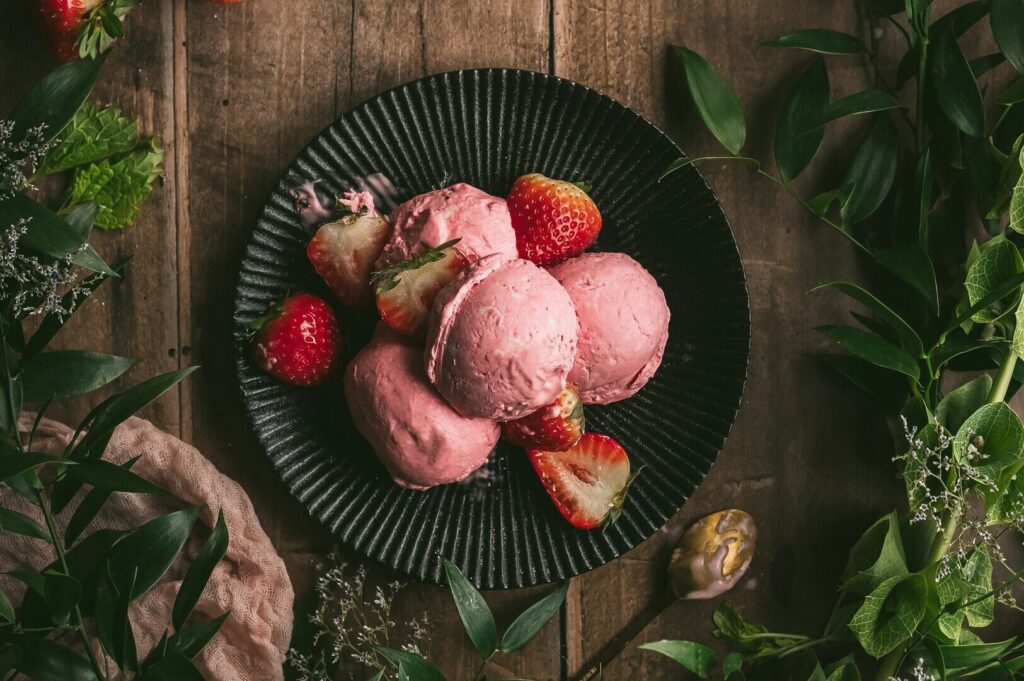 Strawberry ice cream served on a black plate, surrounded by green leaves and delicate flowers, showcasing food photography.