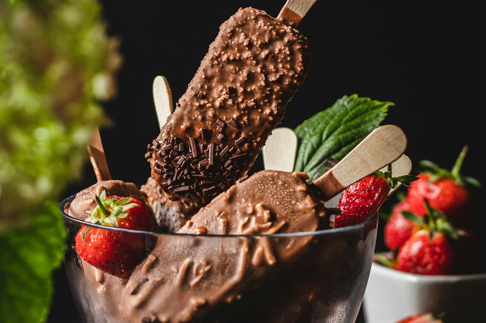 Chocolate-covered ice cream bars with nuts and sprinkles in a glass bowl, artfully arranged for food styling photography, garnished with strawberries and mint leaves, with more strawberries in the background.