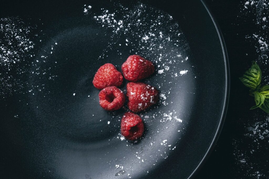 Raspberries on a black plate, garnished with sugar and fresh mint leaves, showcasing vibrant food art photography.