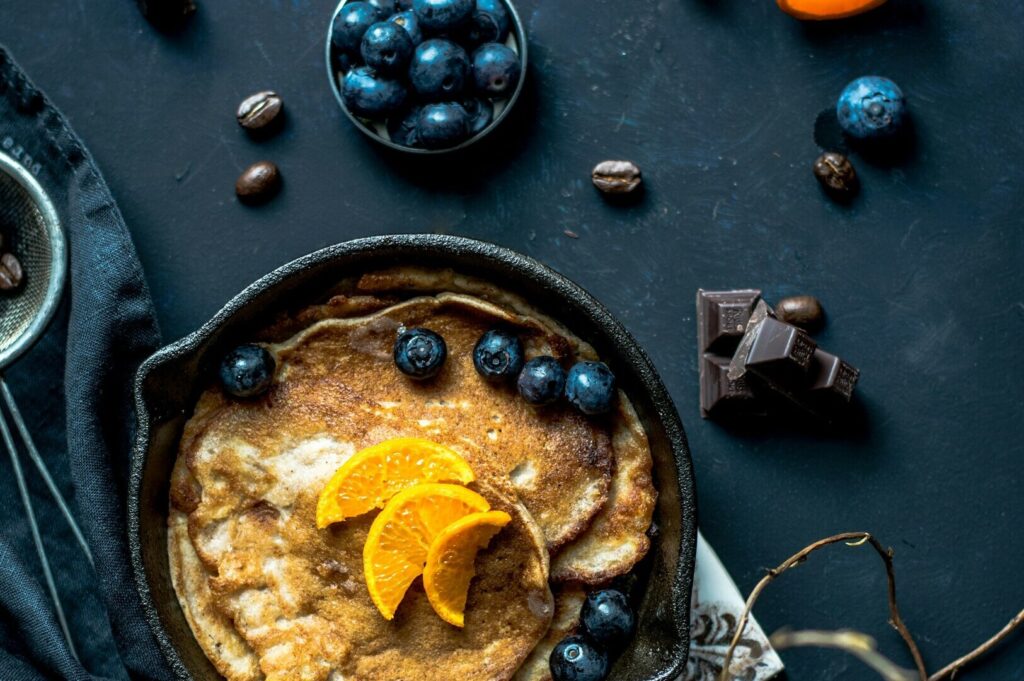 A pan filled with fluffy pancakes garnished with fresh blueberries and melted chocolate, set on a dark surface.