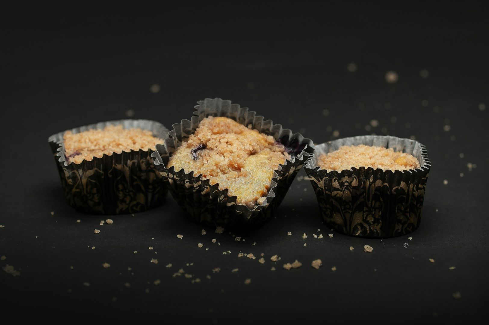 Three blueberry muffins topped with crumble are placed in patterned paper cups against a dark background. Crumbs are scattered around, adding texture.