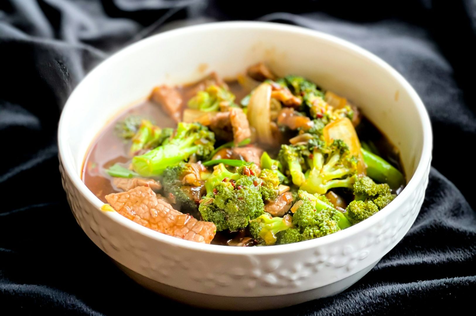 A bowl of beef and broccoli stir-fry on a dark cloth. The dish features tender beef slices and vibrant broccoli in a glossy, savory sauce.