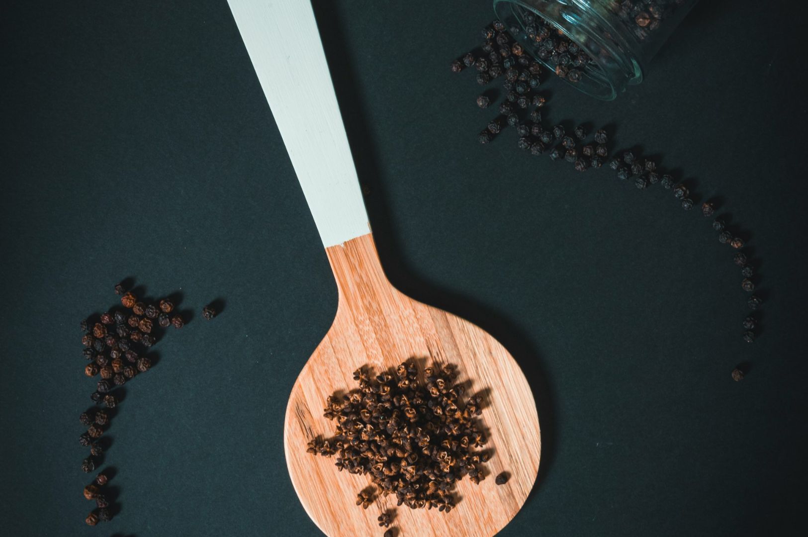A wooden spoon filled with black peppercorns sits on a dark background. Nearby, more peppercorns spill out from a small glass jar onto the surface.