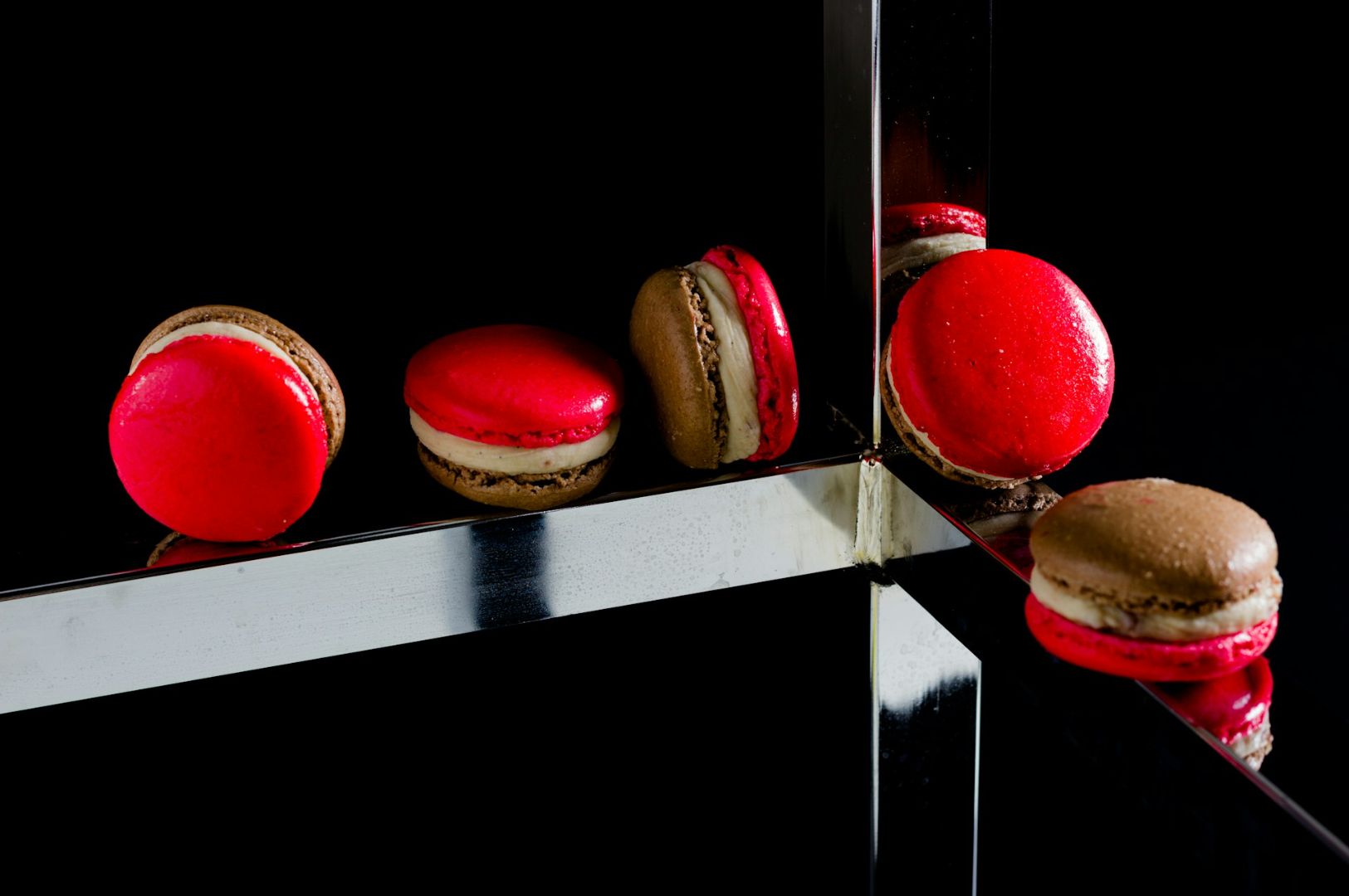 Five vibrant macarons, red and brown, are artfully arranged on reflective metal surfaces against a black background, creating a chic, modern vibe.