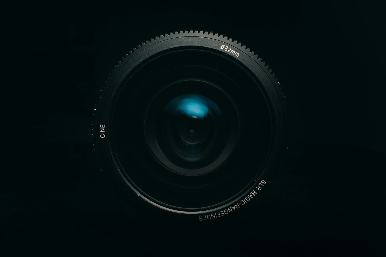 Close-up of a camera lens against a dark background. The glass reflects a soft blue light, conveying a mysterious and professional tone.