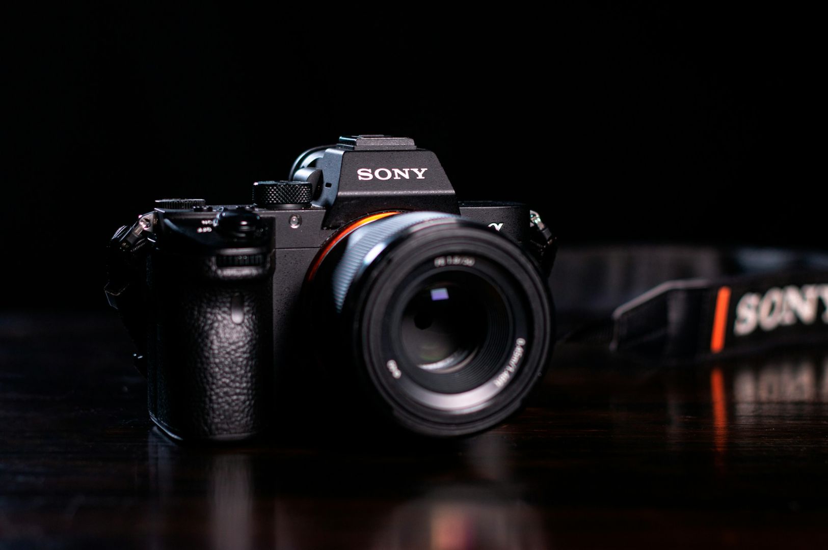 A black Sony camera with a lens sits on a dark wooden surface against a black background. The attached camera strap is partially visible to the right.