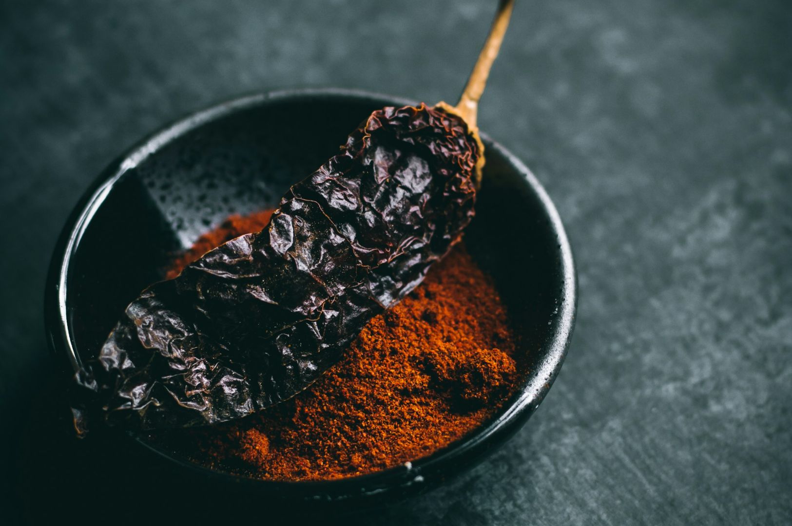 A dark, wrinkled chili pepper rests on vibrant red chili powder in a black bowl, set against a dark, textured background, evoking a spicy and earthy tone.