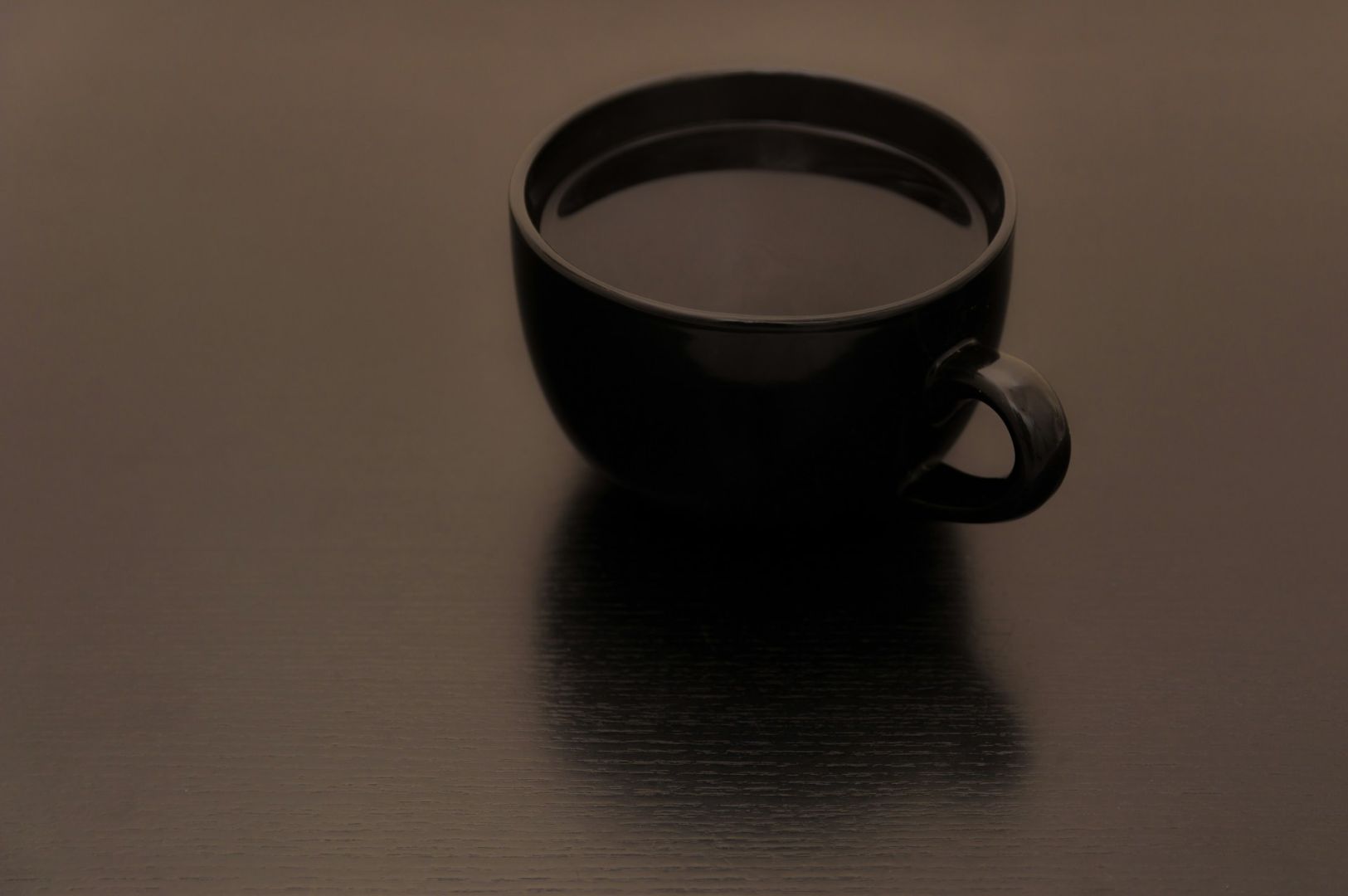 Black ceramic cup filled with dark coffee sits on a smooth, reflective wooden table. The moody lighting creates a calm, minimalist atmosphere.
