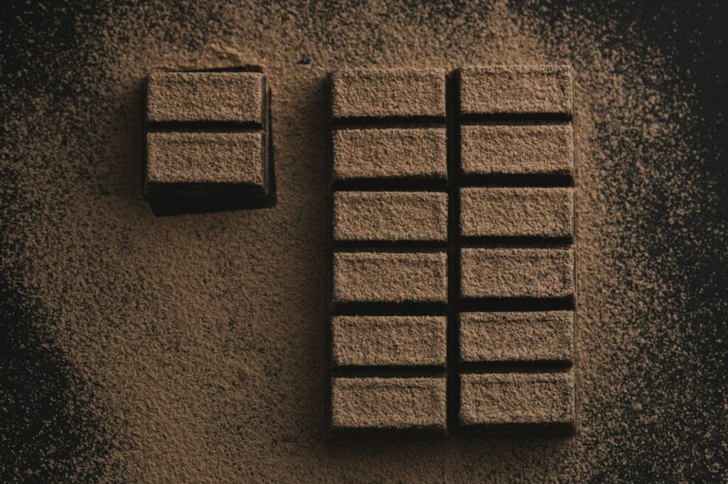 Two sets of chocolate bars are dusted with cocoa powder on a dark surface. The left block is smaller with four pieces, and the right is larger with twelve pieces.