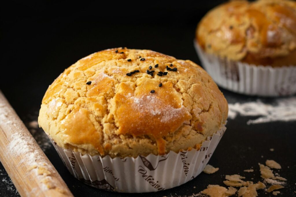 Close-up of a golden, crusty muffin in a white paper cup, dusted with flour and black sesame seeds. A wooden rolling pin lies nearby on a dark surface.