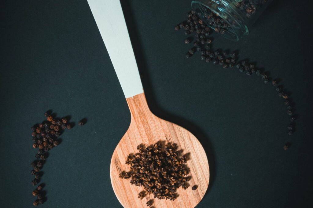 Wooden spoon with peppercorns on a dark surface. A glass jar lies tipped over, spilling more peppercorns, creating a rustic, culinary vibe.