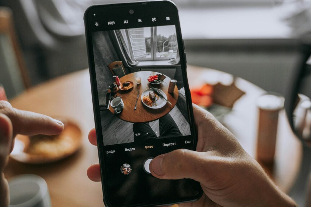 Hands hold a smartphone capturing a wooden table set with plates, cutlery, and food. A window softly lights the scene, creating a cozy atmosphere.