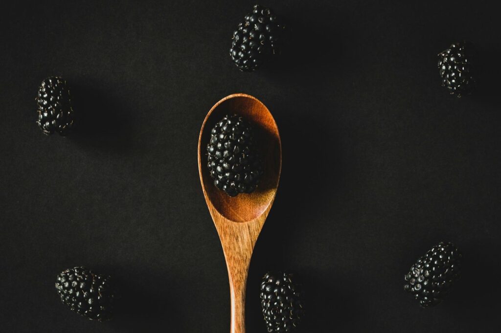 A wooden spoon holding a blackberry is centered on a black background, surrounded by scattered blackberries. The composition is simple and elegant.