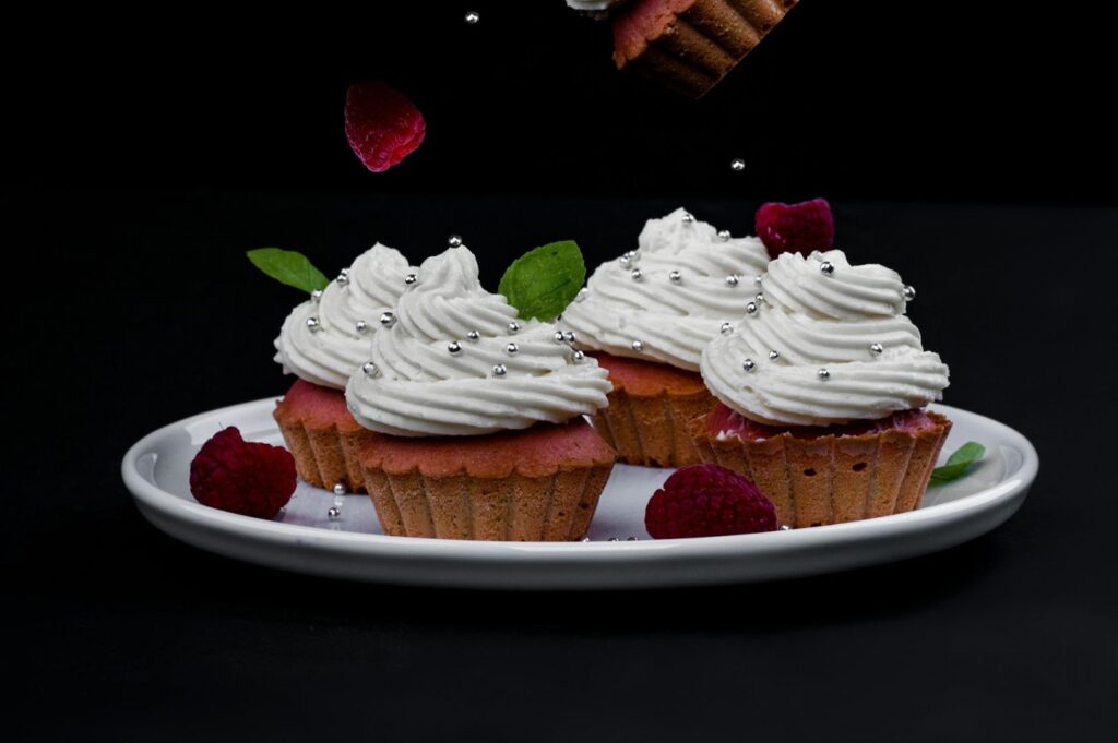 Three cupcakes with swirled white frosting and silver beads sit on a white plate. Raspberries and mint leaves accent the scene against a dark background.