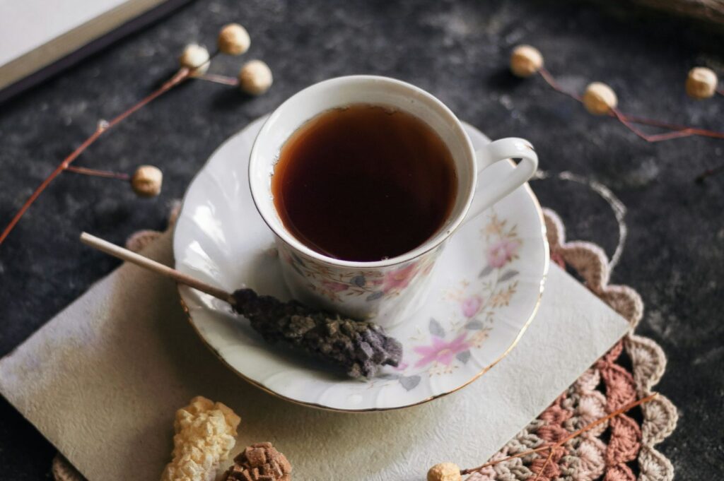 A floral teacup filled with dark tea sits on a matching saucer, next to a stick of sugared lavender. Rustic decor and soft lighting evoke a cozy, vintage ambiance.