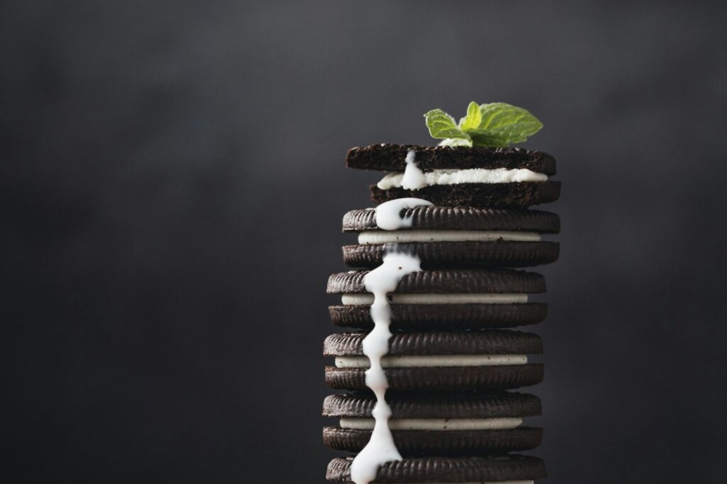 A tall stack of chocolate sandwich cookies with white cream filling is topped with mint leaves and drizzled with milk on a dark background. Rich and indulgent.
