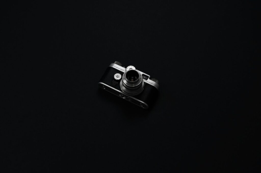 A small, vintage black camera with a silver lens is centered on a dark black background, emphasizing its retro design and sleek form.