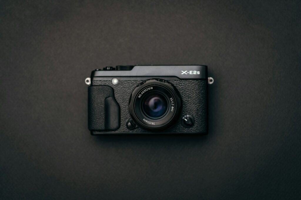 A black X-E2S camera sits centered on a dark, textured surface. The lens reflects subtle light, highlighting a sleek, minimalist design.