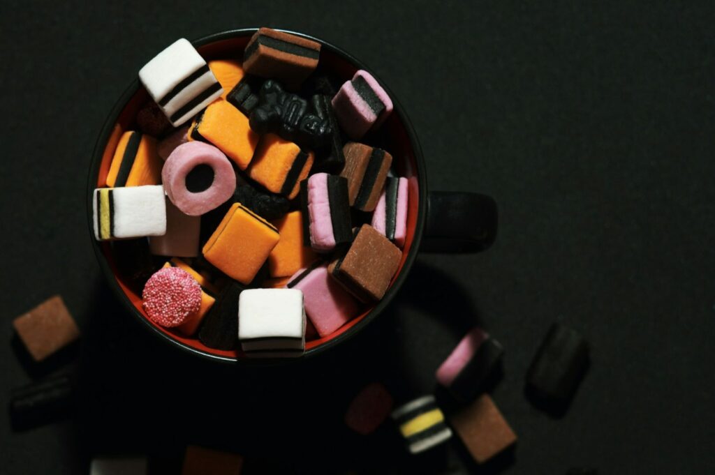 A black cup filled with brightly colored licorice allsorts candies in orange, pink, white, and brown hues on a dark background. Some candies are scattered around.