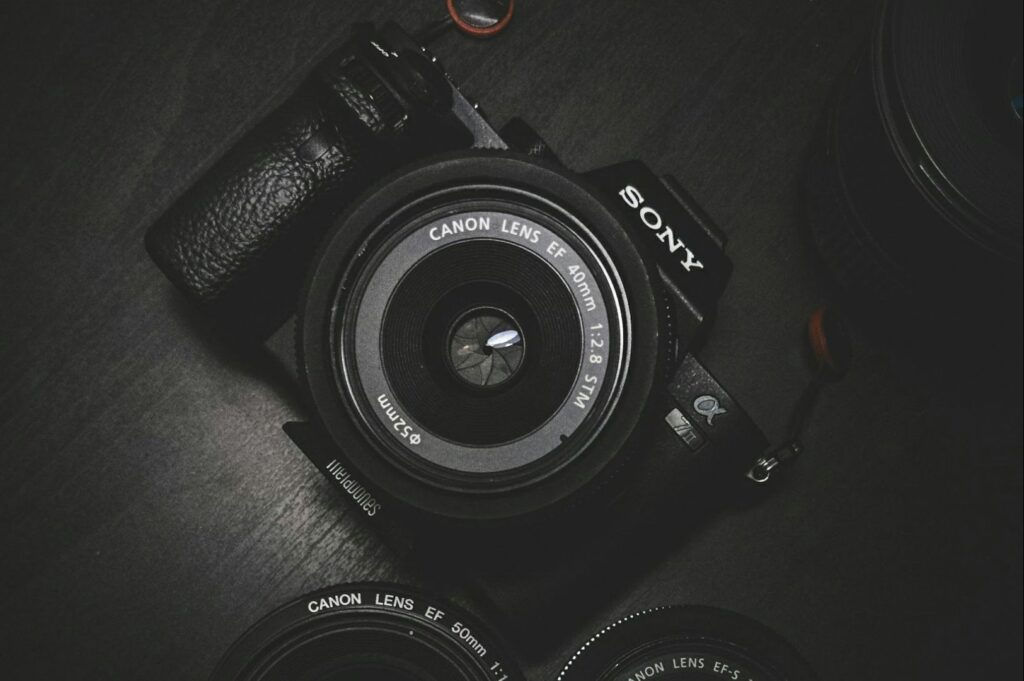 A Sony camera with a Canon lens attached, surrounded by additional lenses on a dark surface. The image conveys a mood of professional photography.