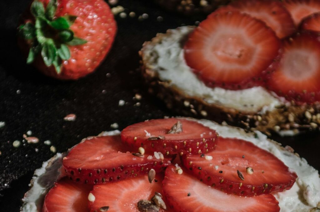 Close-up of sliced strawberries on creamy toast, sprinkled with seeds, against a dark background. Nearby, a whole strawberry adds freshness.