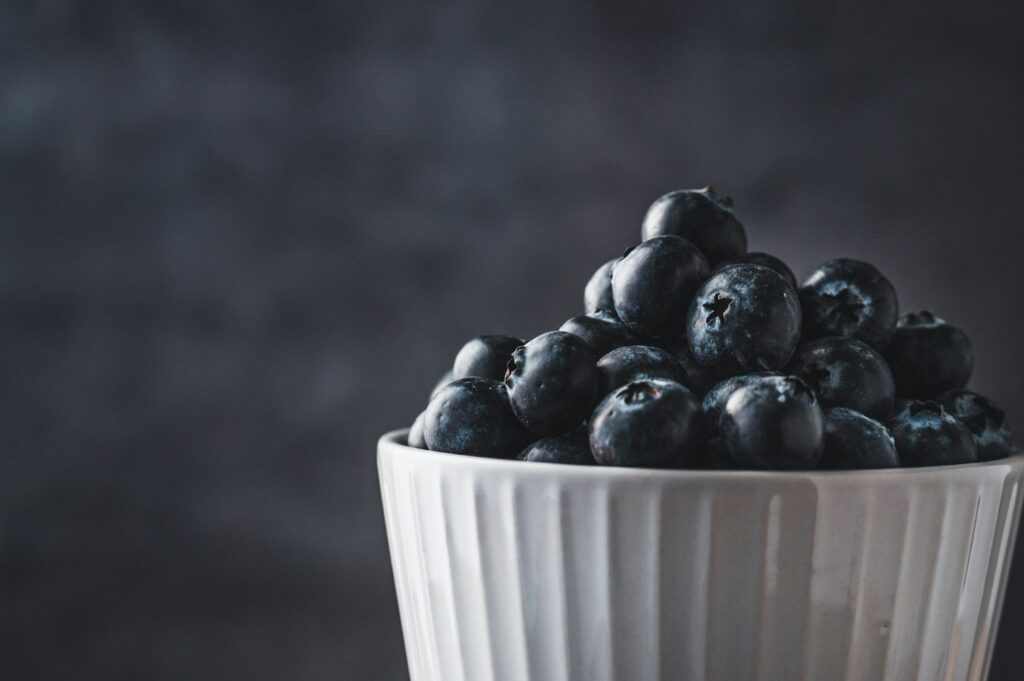 A white ceramic bowl filled with fresh blueberries against a dark, blurred background. The image conveys a fresh, natural, and healthy tone.