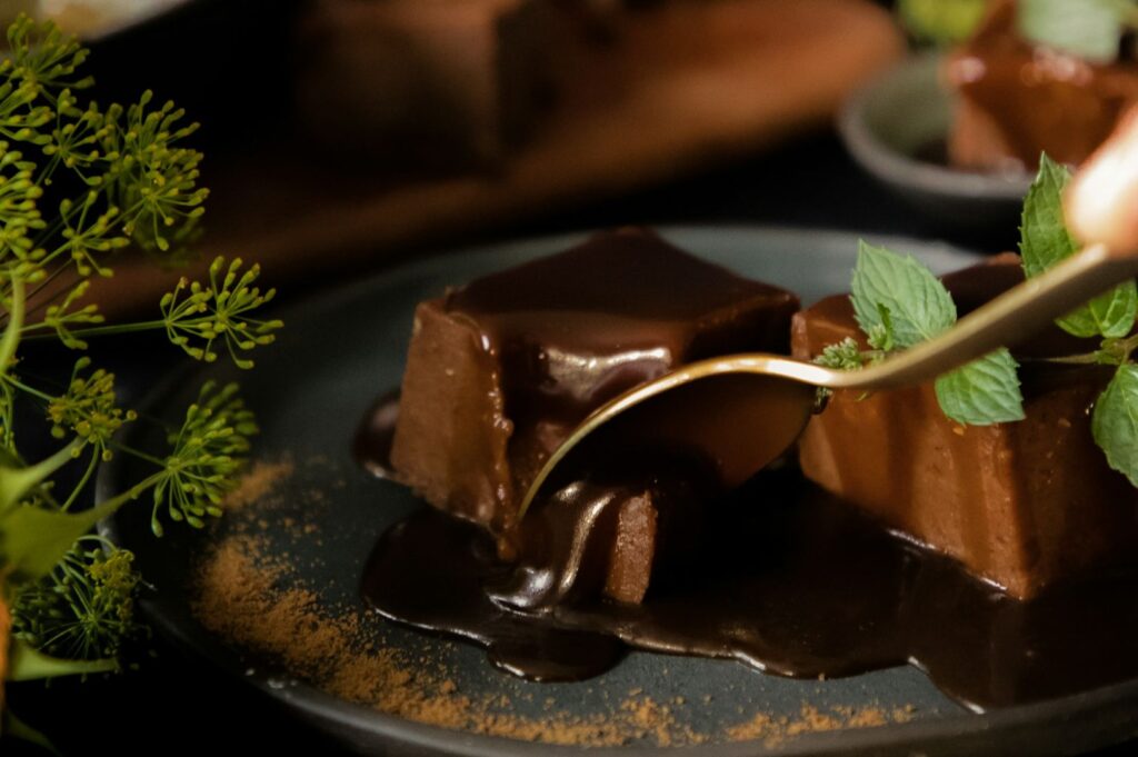 Two rich chocolate desserts topped with glossy sauce on a gray plate. A gold spoon is poised to take a bite. Mint leaves add a fresh touch nearby.