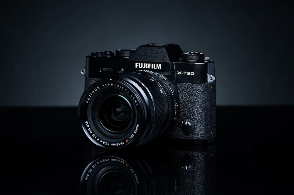 A black Fujifilm X-T30 camera with a Fujinon lens, placed on a reflective surface. The lighting highlights the camera's textures and details.