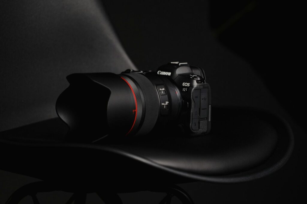 A Canon EOS camera with a large lens, featuring a red ring, rests on a black chair. The setting is dimly lit, creating a sleek, professional tone.