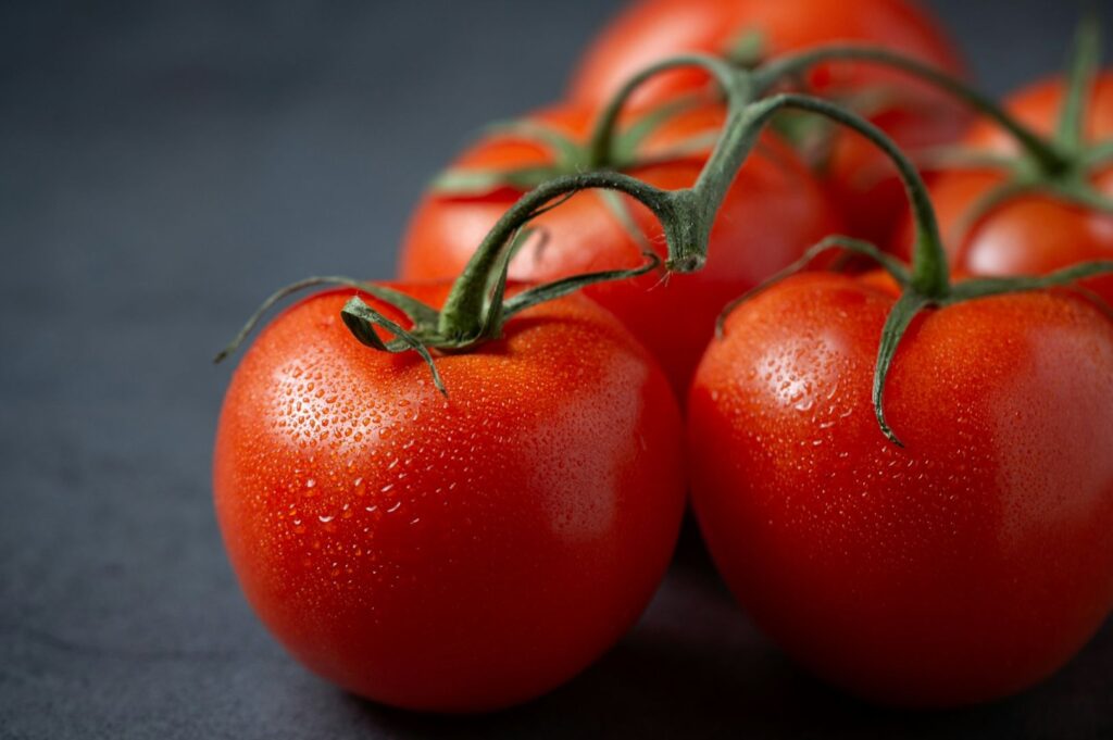 Close-up of vibrant red tomatoes with green stems, featuring a fresh, dewy texture. They rest on a dark surface, conveying freshness and vitality.