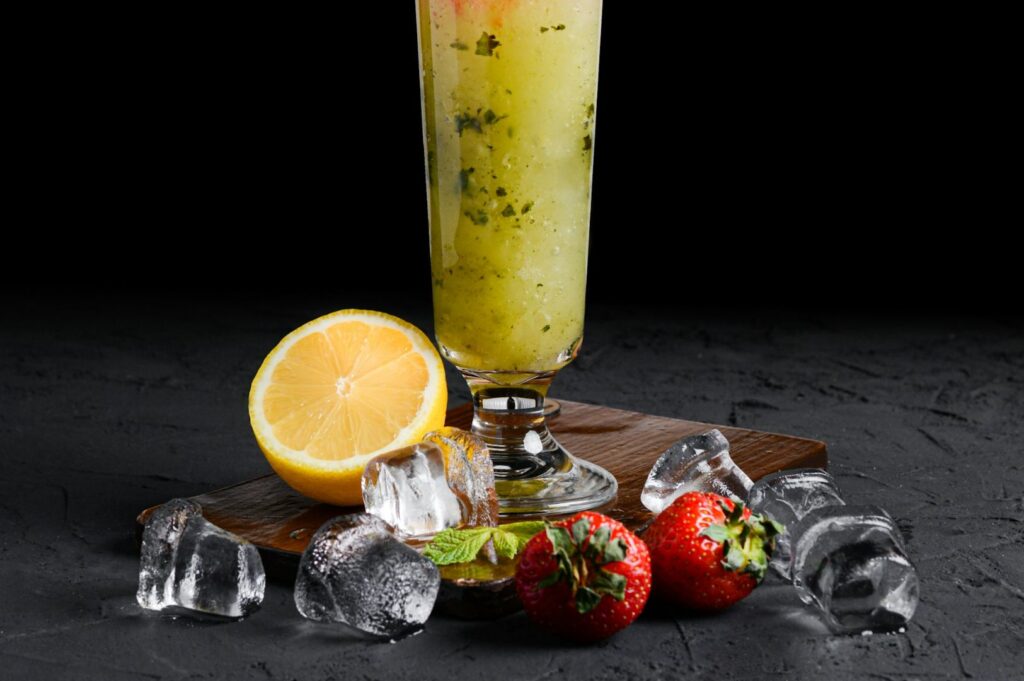 A tall glass of refreshing lemon mint drink on a wooden board, accompanied by fresh strawberries, lemon half, mint leaves, and scattered ice cubes.