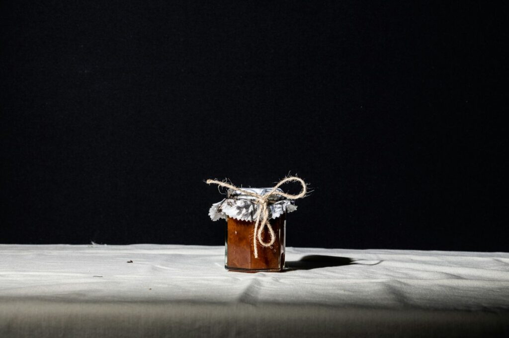 Small jar of jam with lace-covered lid tied with twine, centered on a white cloth against a dark background. Minimalist and rustic tone.