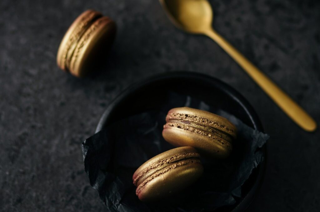 Elegant gold macarons in a black dish on a dark textured surface, paired with a matching gold spoon. The mood is luxurious and sophisticated.