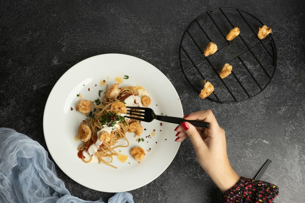 A hand with red-painted nails holds a fork poised over a white plate of shrimp pasta with herbs and sauce. Nearby, a grill rack holds more shrimp.
