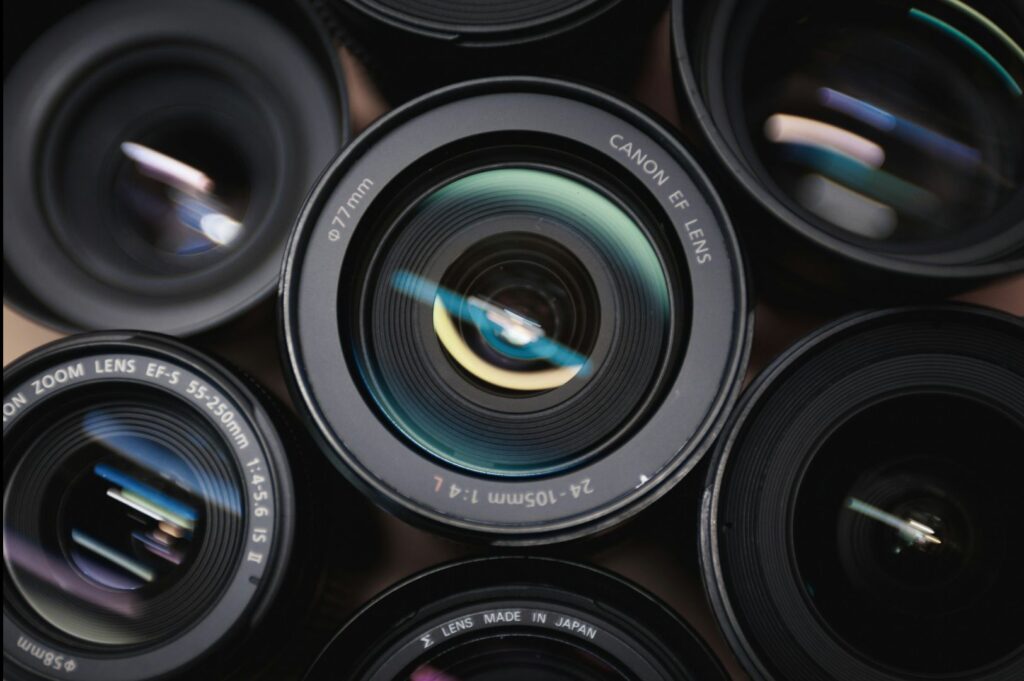 Close-up shot of multiple camera lenses with colorful reflections. Central lens labeled "Canon EF" draws focus. Mood is technical and artistic.