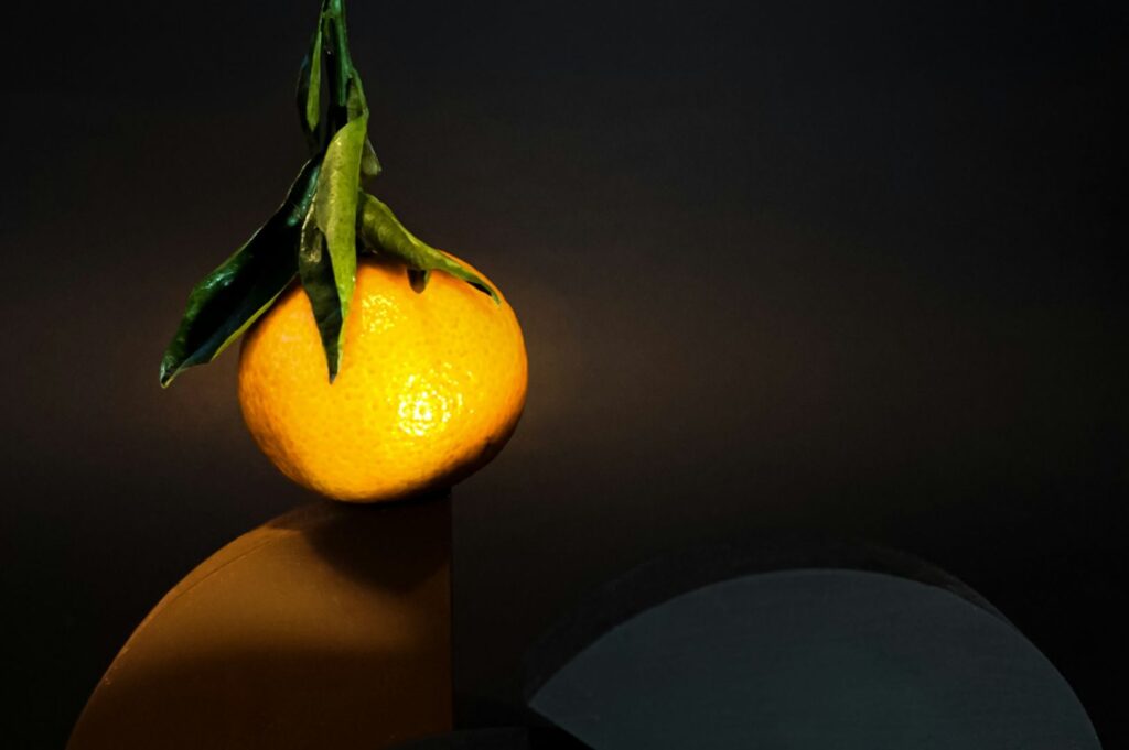 A ripe orange with green leaves rests on a curved, abstract surface against a dark backdrop. The lighting creates a dramatic, artistic tone.